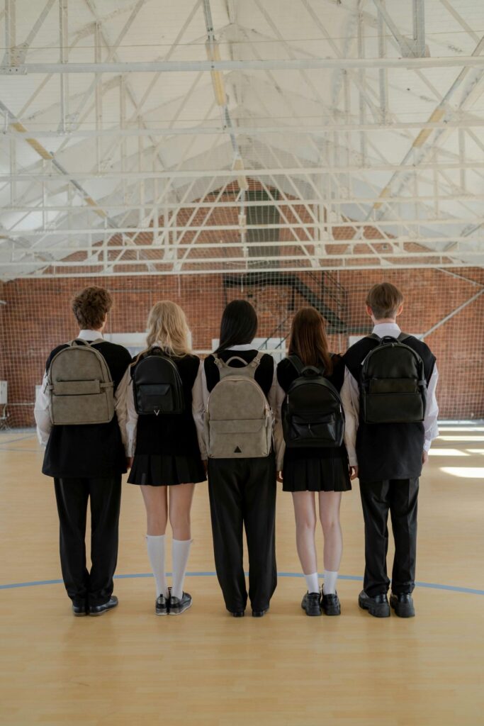 Fünf Schülerinnen und Schüler einer Secondary School in den USA stehen mit Schuluniformen und Rucksäcken in einer Turnhalle und blicken nach vorn.
